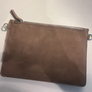 Beige mocka kuvertväska med dragkedja - Stilren beige kuvertväska från Carin Wester i mjuk mocka. Väskan har silverfärgad dragkedja och minimalistisk design. Perfekt storlek för det viktigaste och har en innerficka med dragkedja. Enkel att matcha till olika outfits.