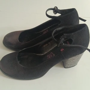 Svarta mockapumps med mönstrad klack - Snygga svarta pumps i mocka från Naf Naf med rund tå och knytband runt vristen. Klacken är medelbred och har ett coolt svartvitt mönster som sticker ut. Perfekta för dig som vill ha något unikt och trendigt till outfiten. Snöret till ena klacken är lite stretchad men går bra att använda ändå. 