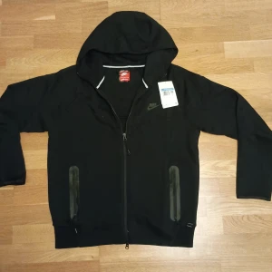 Svart Nike tech plus short - Svarta shorts från Nike med diskret logga på vänster ben. De har elastisk midja med snörning och dragkedjeficka på sidan. Perfekta för träning eller chill, tillverkade i mjuk bomullsblandning för maximal komfort.