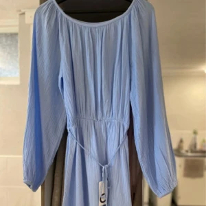 Ljusblå långärmad klänning med knyt - Superfin ljusblå klänning med långa ärmar och knyt i midjan. Den har en avslappnad passform och är gjord i ett lätt, skrynkligt bomullstyg. Perfekt för varma dagar när du vill känna dig både bekväm och snygg. Klänningen slutar ungefär i mitten på låret. 