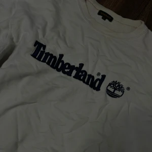 Vit Timberland tröja med broderad logga - Vit tröja från Timberland med stor, broderad blå logga och text på bröstet. Klassisk rund halsringning och mjukt bomullsmaterial. Perfekt för dig som gillar streetwear och vill ha en clean look med tydlig branding.