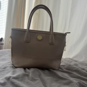 Beige handväska från Don Donna - Snygg beige handväska från Don Donna med guldfärgade detaljer och logga framtill. Väskan har två rejäla handtag, dragkedja upptill och extra fack med dragkedja på baksidan. Tillverkad i skinnimitation med stilren och klassisk design.