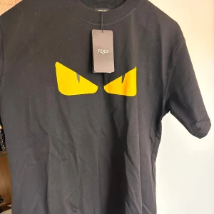 Svart Fendi t-shirt med gula ögon - Cool svart t-shirt från Fendi med ikoniska gula ögon framtill. Klassisk rund hals och korta ärmar. Tillverkad i mjuk bomull som känns skön mot huden. Perfekt för dig som vill sticka ut med en stilren men edgy look.