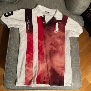 Randig pikétröja från Ralph Lauren M - Snygg pikétröja från Ralph Lauren i storlek M. Tröjan har breda vertikala ränder i vinrött, rött och vitt framtill, stor logga på bröstet och broderad text på ena ärmen. Klassisk krage och knappar framtill. Perfekt för en sportig och stilren look.