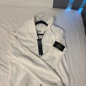 Vit hoodie från Stone Island med dragkedja - Vit hoodie från Stone Island med klassisk logga på ärmen. Tröjan har huva, hel dragkedja framtill och är tillverkad i mjuk bomull. 