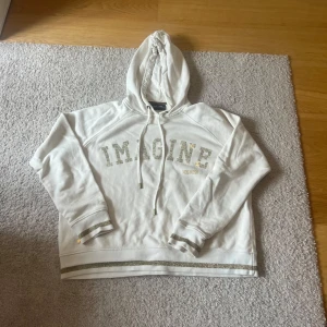 Vit hoodie med glitterdetaljer Mos Mosh - Vit hoodie från Mos Mosh med texten 'IMAGINE' i glittriga bokstäver på bröstet. Tröjan har huva med snörning och silvriga glitterränder vid ärmslut och nederkant. Perfekt för dig som gillar en chill och snygg stil.