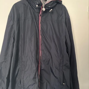 Mörkblå vindjacka från Moncler - Snygg svart vindjacka från Moncler med huva och dragkedja med rödvit detalj. Jackan har klassisk Moncler-logga på ärmen och praktiska fickor med dragkedja. Storlek L men liten så passar M/L
