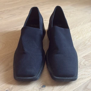 Svarta loafers från Vagabond - Stilrena svarta loafers från Vagabond med fyrkantig tå och minimalistisk design. Skorna har en platt sula och är tillverkade i textil med insida i syntet. Perfekta för dig som gillar en clean och modern look.