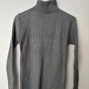 Grå ribbad polotröja - Snygg grå polotröja med ribbat mönster och lång ärm. Tröjan har en hög krage som ger en clean look och är tillverkad i ett mjukt, stickat material. Perfekt att styla med jeans eller kjol för en chill vibe.