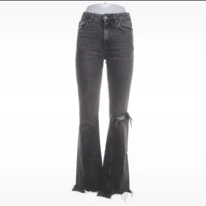 Svarta bootcut jeans med slitningar - Snygga svarta bootcut jeans med mid midja och slitna detaljer vid knät och råa benslut. Jeansen har klassisk femficksdesign och dragkedja. Perfekta för dig som gillar en stockholm och trendig look.