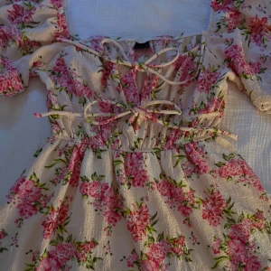 Blommig kortklänning med snörning - Supersöt kortklänning i ljusbeige med rosa blommor och gröna blad. Klänningen har puffiga korta ärmar, smock i midjan och snörning framtill. Perfekt för sommarens alla tillfällen.