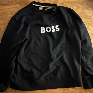 Svart sweatshirt från BOSS - Svart sweatshirt från BOSS med vit logotyp tryckt på bröstet. Klassisk rund halsringning och långärmad modell. Tillverkad i mjuk bomull som känns skön mot huden. Perfekt för en clean och stilren look. Helt ny använd 1 gång , köpt från room 1006 för 1300kr 