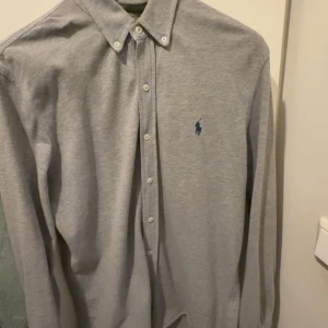 Grå skjorta från Polo Ralph Lauren - Snygg grå skjorta från Polo Ralph Lauren i bomullsmaterial. Klassisk button-down krage, långa ärmar och broderad blå logga på bröstet. Skjortan har knäppning framtill och passar perfekt till en clean och stilren look.