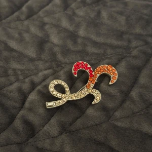 brosch med strass i rött och orange - Unik brosch i silvrig metall, dekorerad med gnistrande strass i rött, orange och klara stenar. Snygg, böljande form som sticker ut och ger en cool touch till din outfit. Perfekt accessoar för dig som gillar färg och bling.
