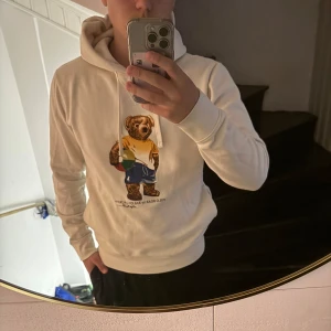 Vit hoodie Polo Bear Ralph Lauren M - Vit hoodie från Polo Ralph Lauren med ikoniska Polo Bear-trycket på bröstet. Klassisk huva med snörning och ribbade muddar. Skön passform och perfekt för dig som gillar streetstyle med en preppy twist. Har några små fläckar på ena armen som knappt märks