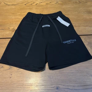 Svarta shorts Essentials Fear of God - Svarta shorts från Essentials Fear of God med elastisk midja och långa snören framtill. Klassisk loose fit och diskret logga på vänster ben. Perfekta för chill dagar eller streetwear. Mjukt bomullsmaterial och bakficka för extra stilpoäng. Pris kan diskuteras.  Längd: 47cm
