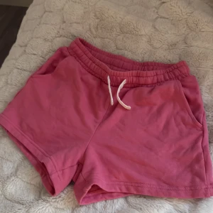 Rosa shorts med snörning från Lindex - Snygga rosa shorts från Lindex med elastisk midja och vit snörning framtill. Mjukt bomullsmaterial och två fickor på sidorna gör dem perfekta för chill dagar eller träning. Passformen är normal och de är riktigt sköna att ha på sig under sommaren.