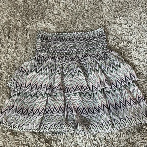 Mönstrad kortkjol från KappAhl - Supersöt kortkjol från KappAhl med färgglatt zickzackmönster i svart, lila, grön och vit. Kjolen har smock i midjan och volangig design som ger en lekfull vibe. Perfekt för dig som gillar att sticka ut med coola mönster och mjuka material.