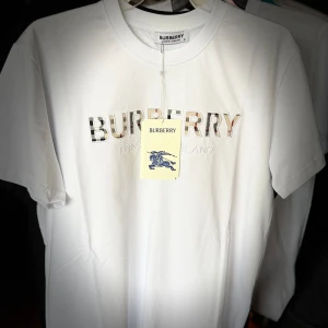 Vit Burberry t-shirt med logga - FINNS I ALLA STORLEKAR!  Snygg vit t-shirt från Burberry med klassisk logga broderad på bröstet i rutigt mönster. T-shirten är i mjuk bomull och har en normal passform med korta ärmar. Perfekt för dig som gillar stilrena och exklusiva plagg.