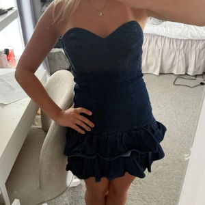 Mörkblå jeansklänning med volang - Supersöt mörkblå jeansklänning med hjärtformad urringning och volangdetaljer nertill. Axelbandslös modell som sitter tight och markerar midjan. Perfekt för dig som vill ha en cool och trendig look med denim vibes. Den är använd fåtal gånger, super söt till sommaren