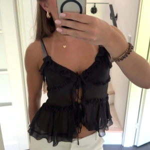 Svart volangig linnetopp från Zara - Supersöt svart linnetopp från Zara med tunna axelband, volanger och knytning framtill. Toppen är i ett lätt, transparent material som ger en luftig känsla och har fina rynkade detaljer längs hela plagget. Perfekt för dig som gillar romantisk och trendig stil.