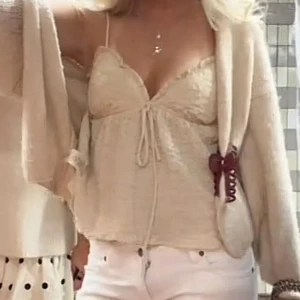 Beige spetsig blus med knyt - Supersöt beige blus med tunna axelband och spetsdetaljer. Toppen har en feminin v-ringning, knyt framtill och lätt volangkant nedtill. Perfekt för dig som gillar romantisk och trendig stil.