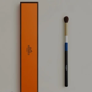Hermès ögonskuggsborste med ask - Lyxig ögonskuggsborste från Hermès med svart, blå och vit skaft, guldfärgad metall och mjuka borst. Kommer i ikonisk orange Hermès-ask. Perfekt för att blenda ögonskuggor och skapa snygga ögonlooks.