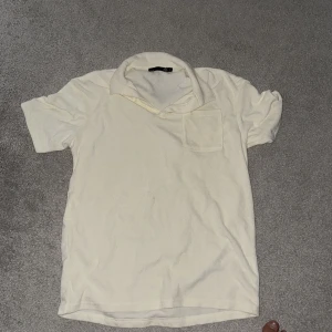 Vit pikétröja från 157, storlek S - Klassisk vit pikétröja från 157 i storlek S. Tröjan har korta ärmar, krage och en bröstficka. Tillverkad i mjuk bomull som känns skön mot huden. Perfekt för en clean och enkel stil.