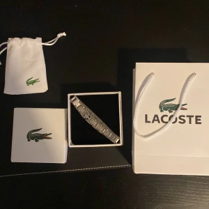 Silverfärgat armband från Lacoste - Stilrent armband från Lacoste i silverfärgad metall med länkdesign och diskret Lacoste-logga på en av länkarna. Kommer i originalförpackning med påse och presentpåse, perfekt som present till någon som gillar klassisk och sportig stil.