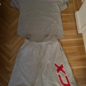 Mjukis set (oversize) - Säljer ett par gråa sweatshorts samt t shirt med snörning i midjan och en stor röd och vit tryckt logga på ena benet. Shortsen har avslappnad passform och är perfekta för chill dagar eller träning. Mjukt bomullsmaterial och sidofickor för extra bekvämlighet.