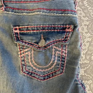 True Religion jeans med färgglada sömmar - Säljer ett par blå jeans från True Religion med ikoniska bakfickor och detaljerade kontrastsömmar i rosa  och vitt. Klassisk femficksmodell, bootcut och dragkedja. Märkets logga på knappar och etikett. Storlek W28 Mått: Innerben-78cm. Midjemått-37cm. Pris kan diskuteras 💕💕💕