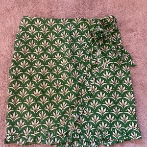 Grön omlottkjol från Urban Threads - Superfin grön kortkjol från Urban Threads med vitt och gult blommönster. Kjolen har omlottmodell med volangkant och en söt rosett i midjan. Perfekt för vår och sommar när du vill sticka ut lite extra!