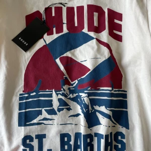 Vit t-shirt med tryck från Rhude - Snygg vit t-shirt från Rhude med stort grafiskt tryck i rött och blått framtill. med klassisk rund hals och korta ärmar. Perfekt statement-plagg för dig som gillar streetwear och coola prints.