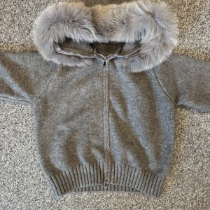 Grå Fur jacket, från alessa  - Mysig grå stickad jacka med dragkedja och huva kantad med fluffig fuskpäls. Jackan har ribbade muddar och en avslappnad passform, perfekt för kyliga dagar. Enkel och stilren design som passar till det mesta.