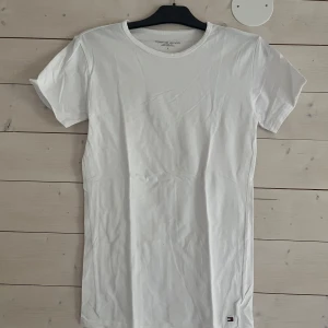 Vit t-shirt från Tommy Hilfiger - Klassisk vit t-shirt från Tommy Hilfiger i mjuk bomull. T-shirten har rund hals, korta ärmar och en liten broderad logga nertill. Perfekt basic-plagg med clean look och skön passform.