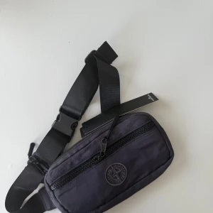 Grå axelväska från Stone Island - Snygg svart axelväska från Stone Island med broderad logga framtill. Väskan har justerbart band, robust dragkedja och ett rymligt huvudfack. 