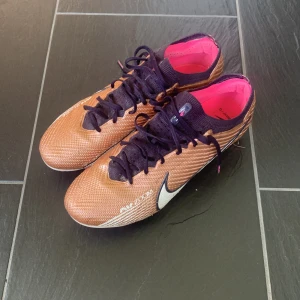  fotbollsskor  - Snygga Nike Mercurial Zoom fotbollsskor i brons med svarta snören och detaljer, samt rosa innersula och dobbar. Don är använda en gång av mig, sen lite mer används av en annan ägare. Men inga problem med skorna förutom att det glansiga har försvunnit lite på sulan vilket brukar hända efter få antal träningar. Annars är skorna helt felfria och kan hålla under en längre tid. Så nästan nyskick! Storlek 42 