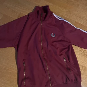 Vinröd Fred Perry kofta - Säljer en vinröd kofta från Fred Perry med vita ränder längs ärmarna och broderad logga på bröstet. Jackan har dragkedja framtill och två fickor med dragkedja.