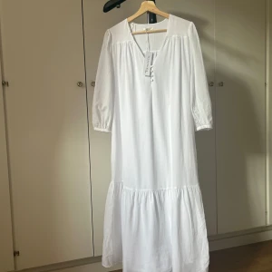 Oanvänd vit långklänning med v-ringning - Luftig vit långklänning från Tamaris med v-ringning, knappar framtill och trekvartsärmar. Klänningen har en volang nertill och är tillverkad i ett lätt material som passar perfekt för varmare dagar.