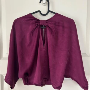 Lila blus med rosett och dragkedja - Säljer en lila blus i glansigt material med lös passform. Blusen har trekvartsärmar, dragkedja nertill och en snygg rosettdetalj i ryggen. Perfekt för dig som gillar unika detaljer och vill sticka ut lite extra.