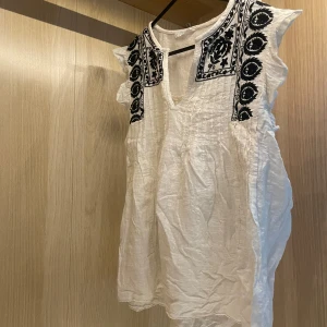 Vit blus med broderi  - Supersöt vit blus från Primark med svarta broderade detaljer vid axlar och halsringning. Blusen är ärmlös med lätt volangkant och har en avslappnad passform. Perfekt för varma dagar och ger en bohemisk vibe.