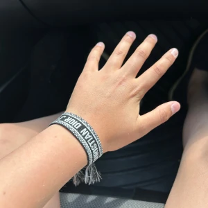 Dior armband  - Snyggt vävt armband från Christian Dior i svart och vitt med texten 'CHRISTIAN DIOR J'ADIOR'. Armbandet har justerbar knytning med tofsar i ändarna och coolt mönster längs kanterna. Perfekt accessoar för att lyfta din outfit.