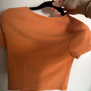 Orange ribbad croppad topp HM - Säljer en orange croppad topp från HM. T-shirten är ribbad med korta ärmar och rund halsringning. Perfekt för dig som gillar en enkel men trendig stil. Materialet är mjukt och stretchigt, vilket gör den superbekväm.