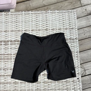 Svarta träningsshorts från SOC - Svarta cykelshorts från SOC med hög midja och tight passform. Perfekta för träning eller sport, tillverkade i stretchigt syntetmaterial som andas. Diskret logga på ena benet. Använda endast under en fotbollsturnering. Storlek 170 men passar xs-s