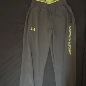 Helt nya svarta träningsbyxor från Under Armour - Svarta träningsbyxor från Under Armour med neongröna loggor och tryck längs benet. Byxorna har elastisk midja och är gjorda i ett lätt syntetmaterial som passar perfekt för sport och träning. Snygg och sportig look med normal passform.
