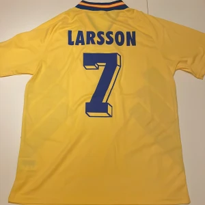 Svenska landslagströja Larsson #7 Adidas - Gul svensk landslagströja replika från Adidas med blå detaljer, Larsson och nummer 7 på ryggen. Klassisk V-ringad krage med blå och gula ränder, stora blå ränder på sidorna och broderat landslagsmärke på bröstet. Tillverkad i lätt och ventilerande polyester. Endast handtvätt gäller