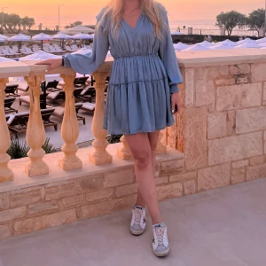 Blå kortklänning från Mango - Supersöt ljusblå kortklänning från Mango med v-ringning, långa ärmar och volangdetaljer vid axlarna. Klänningen har resår i midjan och är tillverkad i ett glansigt, mjukt material som ger en lyxig känsla. Perfekt för sommarens alla tillfällen!