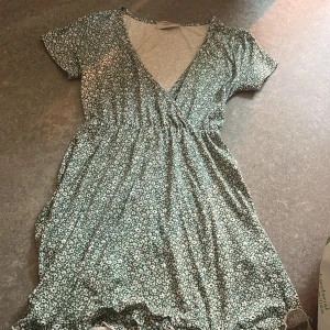 Grön blommig omlottklänning Chiquelle - Supersöt grön kortklänning från Chiquelle med vitt blommönster, omlott och v-ringning. Klänningen har korta ärmar, resår i midjan och volangdetalj nertill. Perfekt för sommarens alla tillfällen! Oanvänd 