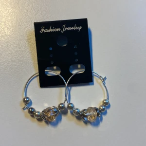 Unika, handgjorda Hoops med pärlor och kristall *Endast 1 Kvar* - Snygga örhängen i hoop-modell med silverfärgade kulor och en större facetterad pärla i beige/brun ton. Perfekta för att ge din look lite extra bling. Lätta att bära och passar till många stilar.