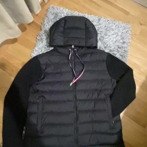 Svart Moncler pufferjacka med huva - Svart pufferjacka från Moncler med huva och coola snören i rött, vitt och blått. Jackan har Moncler-logga på ärmen och dragkedja framtill. Perfekt för dig som vill ha en stilren och varm jacka med streetkänsla.